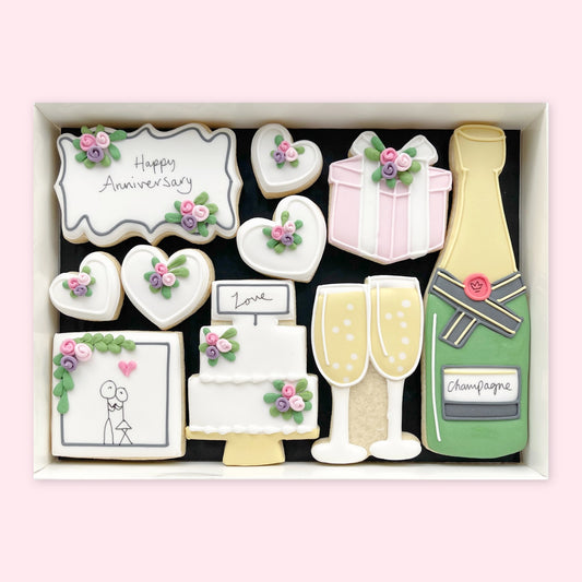 Hand-iced anniversary biscuits in a gift box, featuring a Happy Anniversary plaque, hearts, cake, champagne bottle and glasses with pastel floral details.