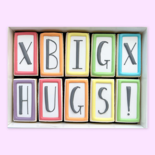 Ten rectangular iced biscuits with colourful borders and black lettering spelling ‘X BIG X’ and ‘HUGS!’, arranged in a white gift box.