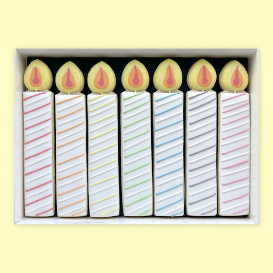 Seven birthday candle-shaped iced biscuits with colourful diagonal stripes and yellow icing flames, arranged in a white gift box.