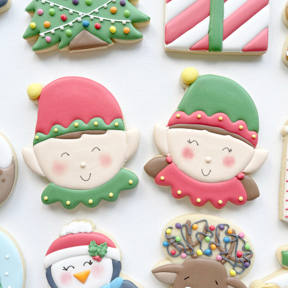 Two smiling elf face biscuits wearing red and green hats, with pointed ears, rosy cheeks and contrasting festive collars.