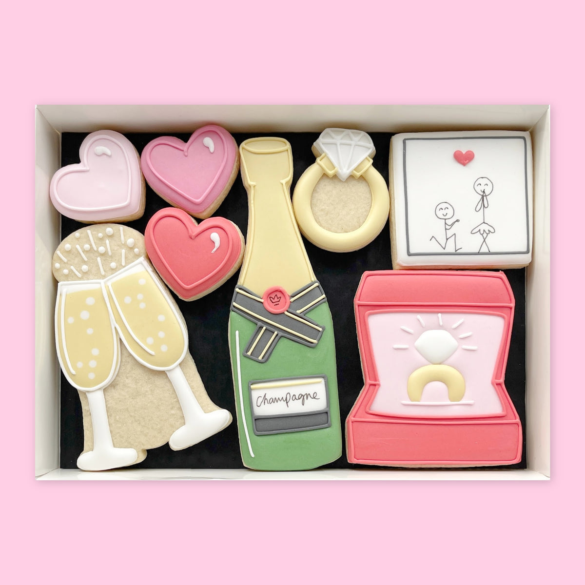 Hand holding a white gift box of engagement-themed iced biscuits, including pink hearts, a diamond ring, champagne designs, ring box and proposal illustration.