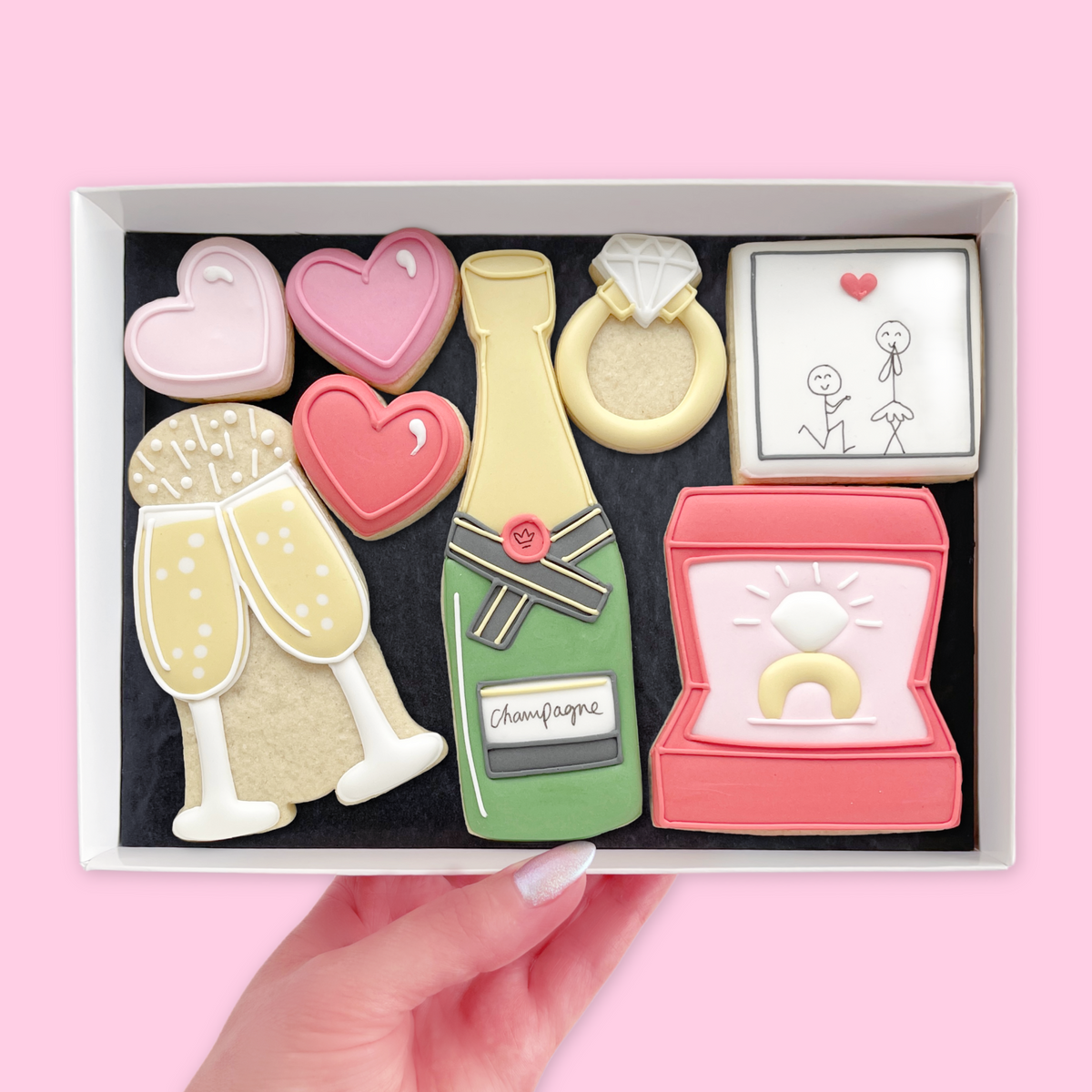 Hand holding a white gift box of engagement-themed iced biscuits, including pink hearts, a diamond ring, champagne designs, ring box and proposal illustration