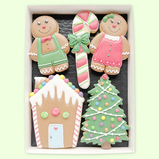 Hand-iced biscuits shaped as a gingerbread boy and girl, a candy cane, a house and a Christmas tree in a white gift box on a pale green background.