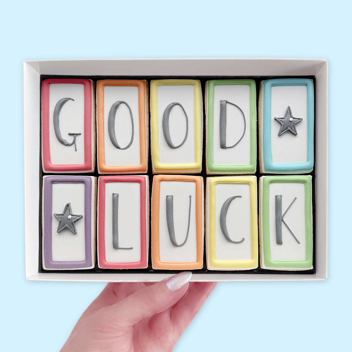 Hand holding a white gift box of ten rectangular iced biscuits with colourful borders, spelling ‘GOOD LUCK’ with two grey star designs