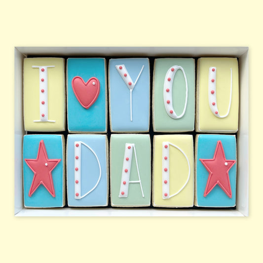 Ten iced biscuits in yellow, blue and green, arranged to read ‘I love you Dad’ with white lettering, a red heart and two red stars in a white gift box.”