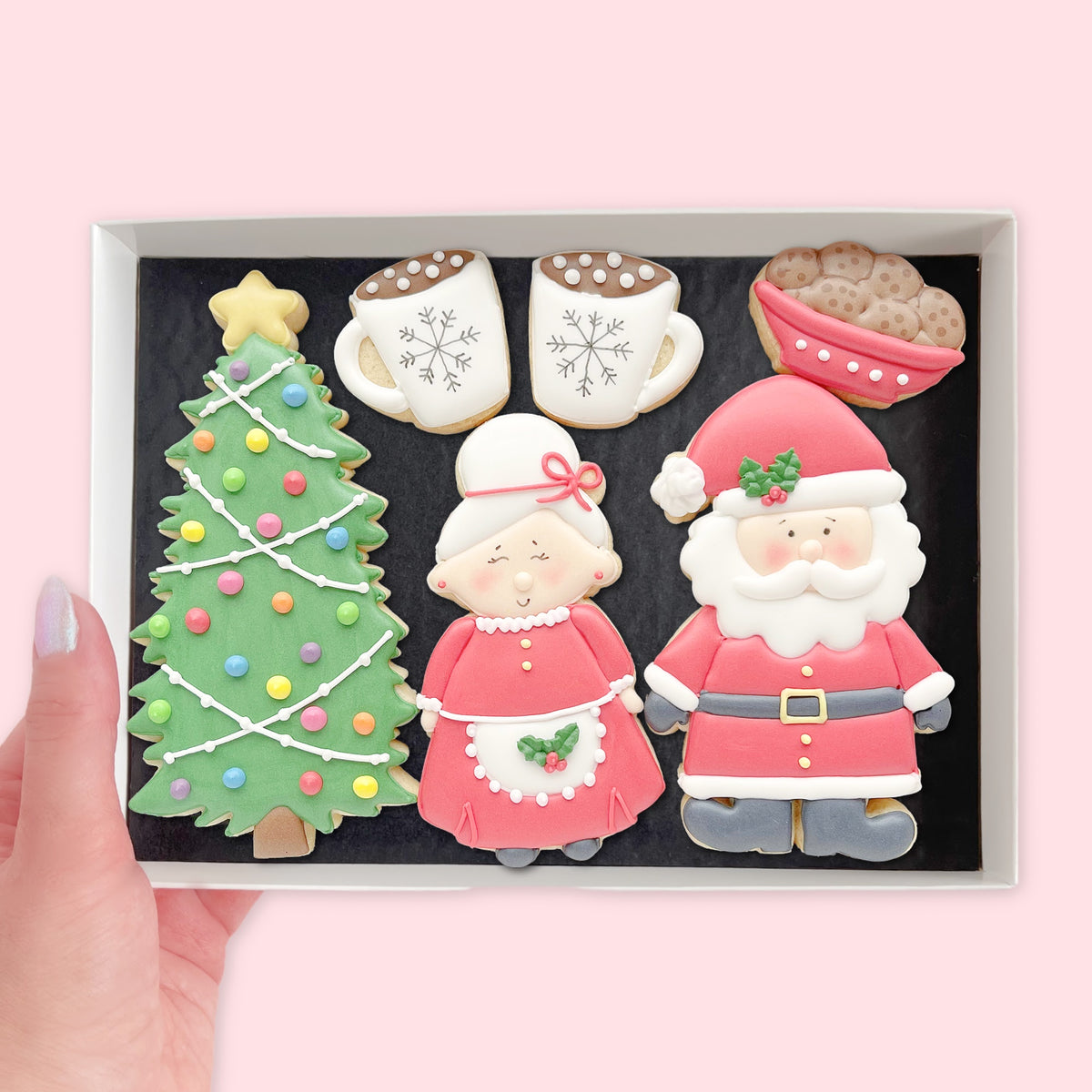 Hand holding a white gift box of hand-iced Mr and Mrs Claus biscuits with a Christmas tree, two hot chocolate mugs and a bowl of biscuits.