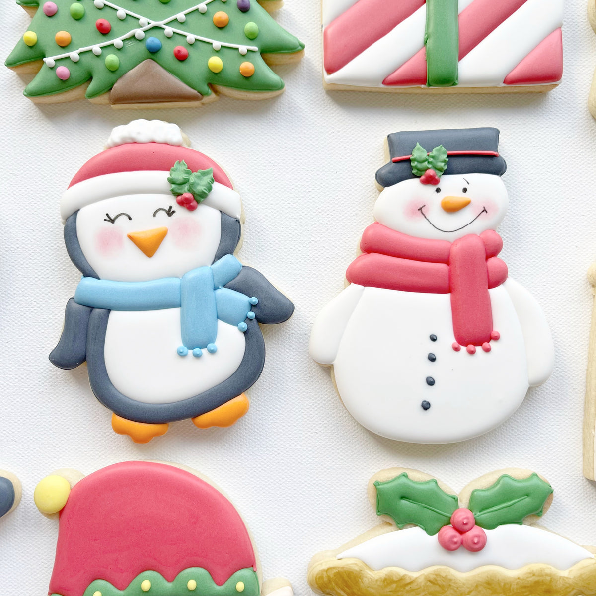 Hand-iced penguin biscuit wearing a red hat and blue scarf beside a snowman biscuit with a black top hat and red scarf.