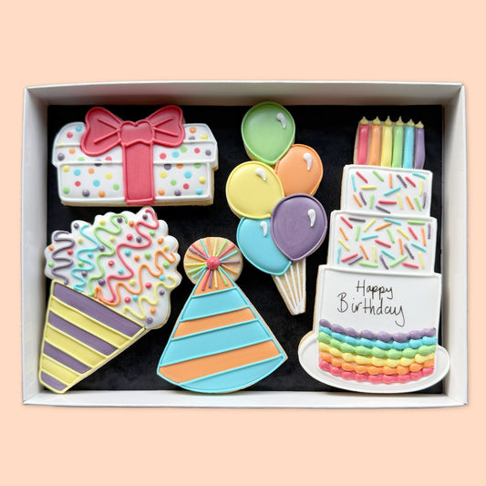 Five colourful hand-iced birthday biscuits shaped as a cake, balloons, gift, party hat and streamer in a white box