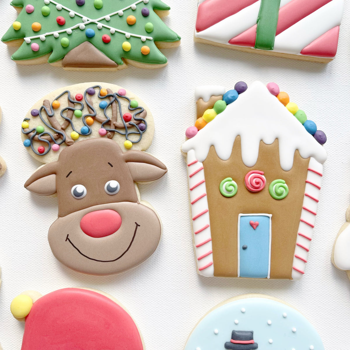 Reindeer face biscuit with a red nose and colourful lights on its antlers beside a house biscuit with a snowy roof and blue door.