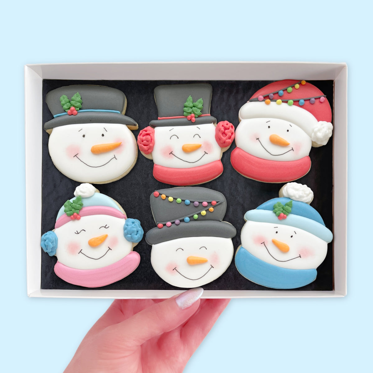 Hand holding an open white gift box containing six hand-iced snowman face biscuits with carrot noses and colourful winter accessories