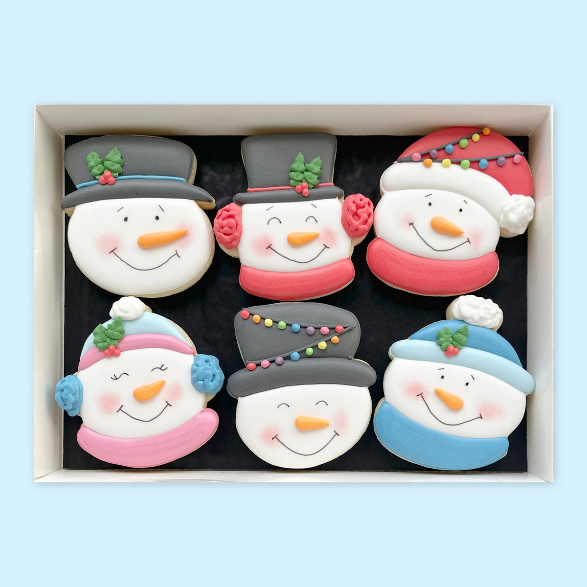 Six hand-iced snowman face biscuits with colourful hats, scarves and earmuffs arranged in a white gift box on a pale blue background.