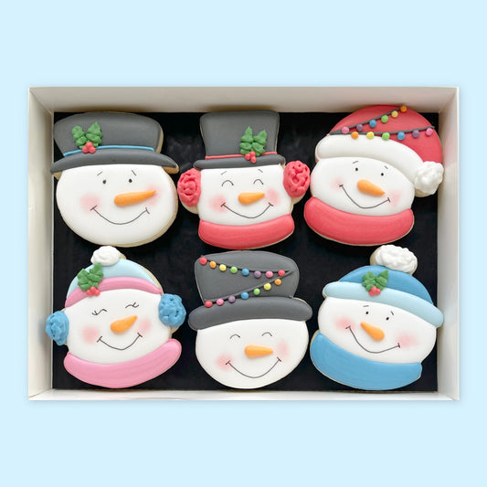 Six hand-iced snowman face biscuits with colourful hats, scarves and earmuffs arranged in a white gift box on a pale blue background.