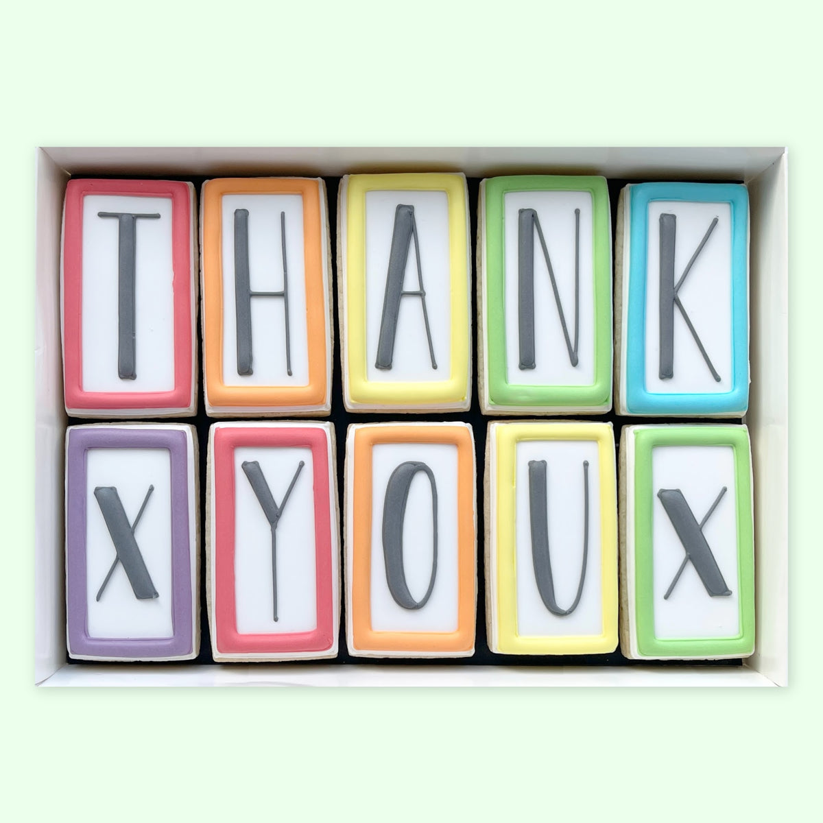 Ten rectangular iced biscuits with colourful borders and black letters spelling ‘THANK X YOU X’, arranged in a white gift box.