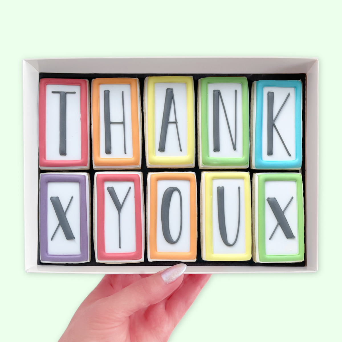 Hand holding a white gift box of ten iced letter biscuits with rainbow-coloured borders, spelling ‘THANK X YOU X