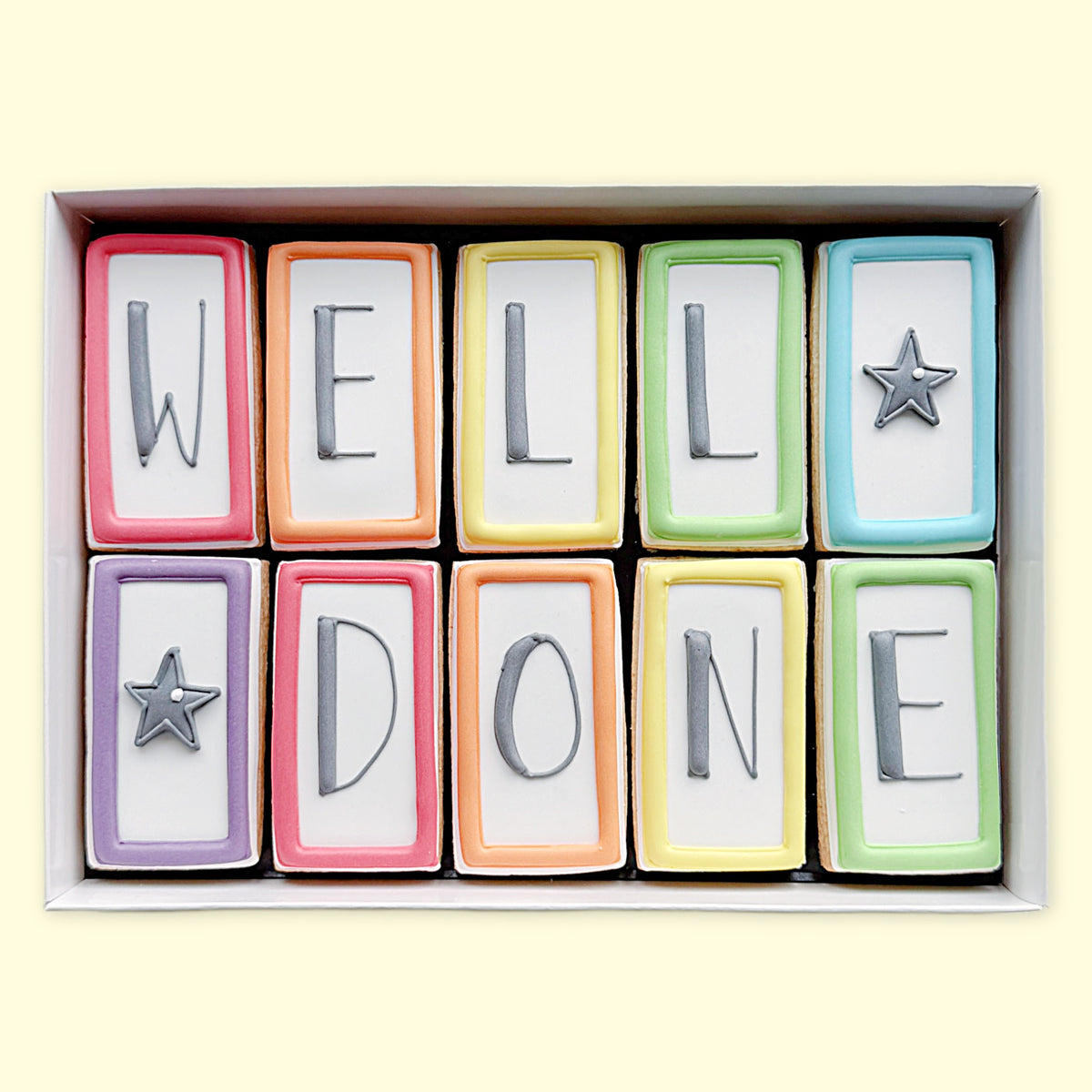 Ten rectangular iced biscuits with colourful borders, grey letters spelling ‘WELL DONE’ and two star designs, arranged in a white gift box.