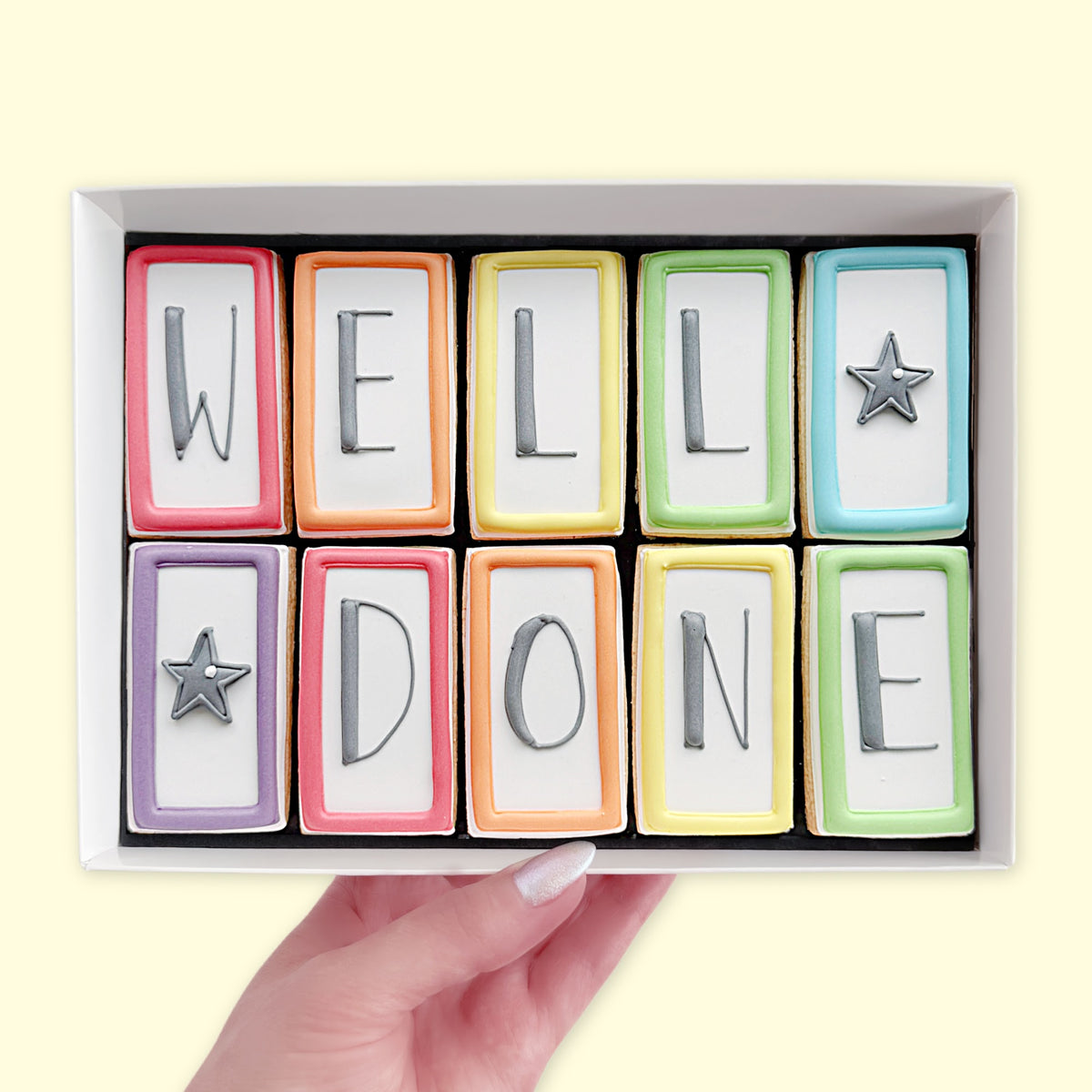 Hand holding a white gift box of ten rectangular iced biscuits with colourful borders, spelling ‘WELL DONE’ with two grey star designs.