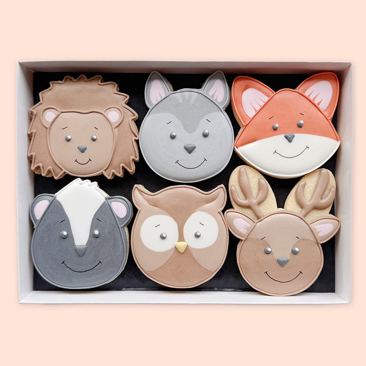 Six hand-iced woodland animal face biscuits arranged in a white gift box on a peach background.