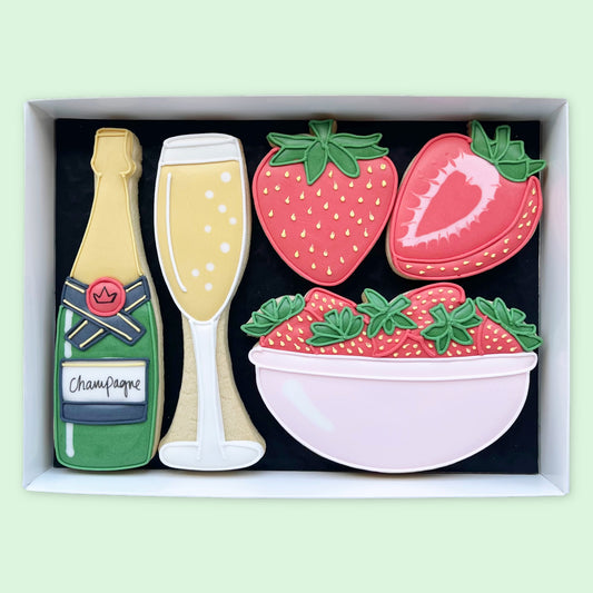 Hand-iced biscuits in a white gift box, featuring a champagne bottle and glass, a whole and halved strawberry, and a pink bowl of strawberries