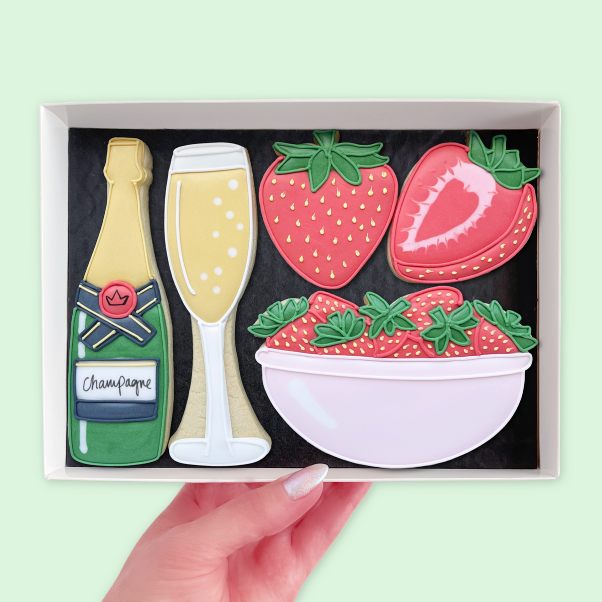 Hand holding a white gift box of iced biscuits shaped as a champagne bottle, champagne glass, strawberries and a pink bowl of strawberries

