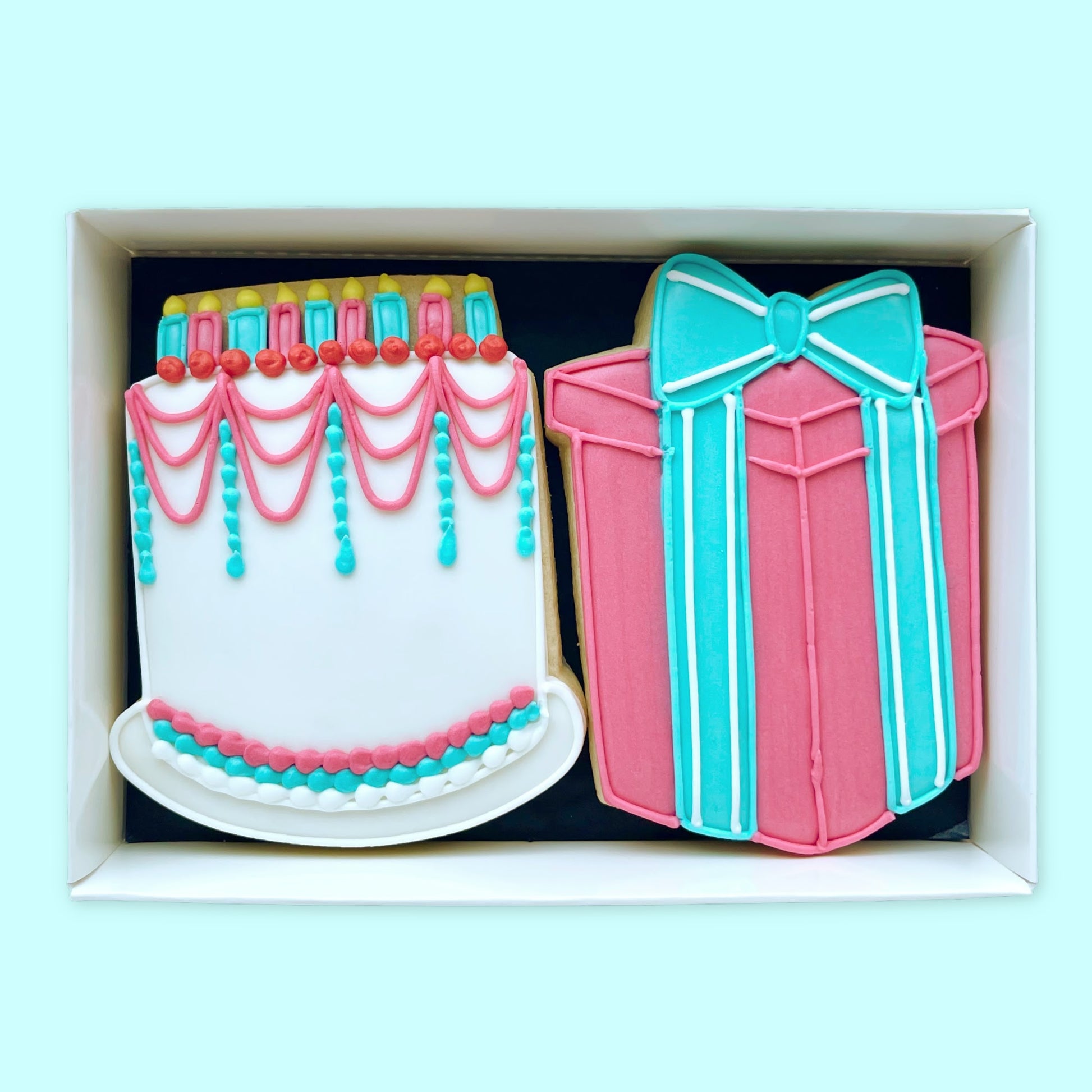 Hand iced Birthday cake with candles and gift with bow biscuits in shades of white blue and pink. in an open white gift box by Katie's biscuit shop