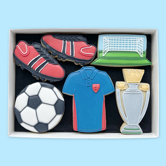Hand-iced football biscuits in a white gift box, featuring red boots, a football, blue shirt, goal on a green pitch and a silver-and-gold trophy.