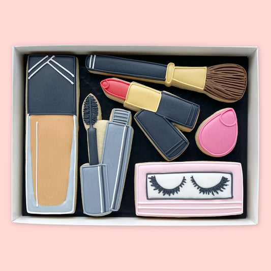 Hand-iced makeup-themed biscuits in a white gift box, featuring foundation, a makeup brush, red lipstick, mascara, a pink sponge and false eyelashes.