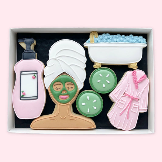 Hand-iced spa-themed biscuits in a white gift box, featuring a woman with a face mask and hair towel, cucumber slices, bubble bath, lotion and pink dressing gown.